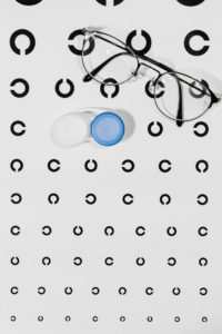 Plakat mit Sehtest auf dem an einer Seite geöffnete Kreise zu sehen sind. Darauf liegen ein Behälter für Kontaktlinsen und eine Brille