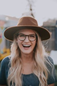 Eine blonde Frau mit braunem Hut und Brille lacht fröhlich