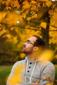 Ein Mann mit Brille steht vor einem Baum mit gelben Herbstblättern