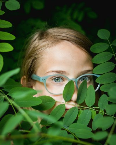 Mädchen mit einer blauen Kinderbrille, das sich hier grünen, kleinen Blättern versteckt